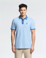 A man wears a Psycho Bunny slim-fit light blue "MENS NEW CASTLE PIQUE POLO - B6K487F200" with an embroidered bunny patch, dark blue collar, and sleeve accents, against a plain background. It's stylishly paired with beige pants.