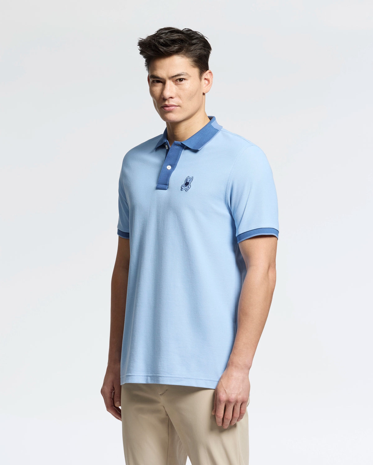 A person is wearing the Psycho Bunny MENS NEW CASTLE PIQUE POLO - B6K487F200, a light blue Pima cotton shirt featuring a slim fit, dark blue collar and cuffs, and an embroidered bunny patch logo on the chest. They're standing against a plain white background.