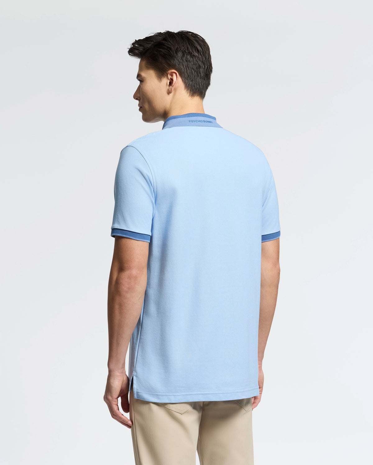 A man wearing a Psycho Bunny's "MENS NEW CASTLE PIQUE POLO - B6K487F200" in slim-fit light blue with dark trim, paired with beige pants, stands against a white background. His short dark hair complements the soft Pima cotton polo featuring an embroidered bunny patch.
