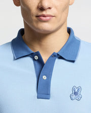 A person showcases the Psycho Bunny MENS NEW CASTLE PIQUE POLO - B6K487F200, a slim-fit, light blue polo made from premium Pima cotton. It features a darker blue collar with two buttons and an embroidered bunny patch with crossed bones below the collar on a plain gray backdrop.