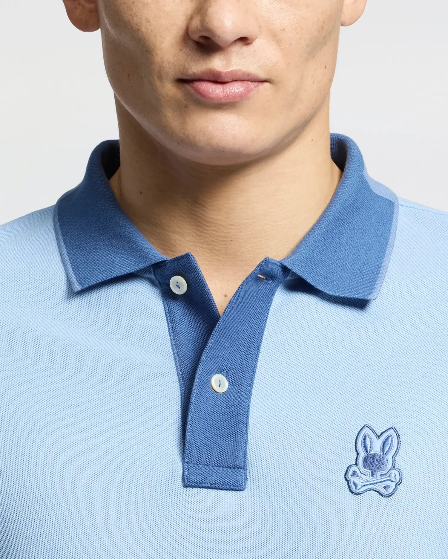 A person showcases the Psycho Bunny MENS NEW CASTLE PIQUE POLO - B6K487F200, a slim-fit, light blue polo made from premium Pima cotton. It features a darker blue collar with two buttons and an embroidered bunny patch with crossed bones below the collar on a plain gray backdrop.