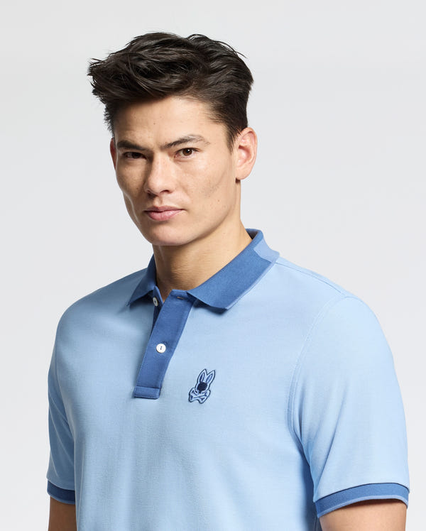A person in an elegant slim fit light blue MENS NEW CASTLE PIQUE POLO - B6K487F200 by Psycho Bunny, with an embroidered Bunny patch on the left chest, made from Pima cotton. They pair it stylishly with their dark hair against a plain background.