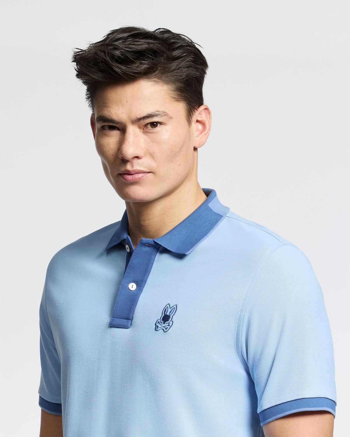 A person in an elegant slim fit light blue MENS NEW CASTLE PIQUE POLO - B6K487F200 by Psycho Bunny, with an embroidered Bunny patch on the left chest, made from Pima cotton. They pair it stylishly with their dark hair against a plain background.