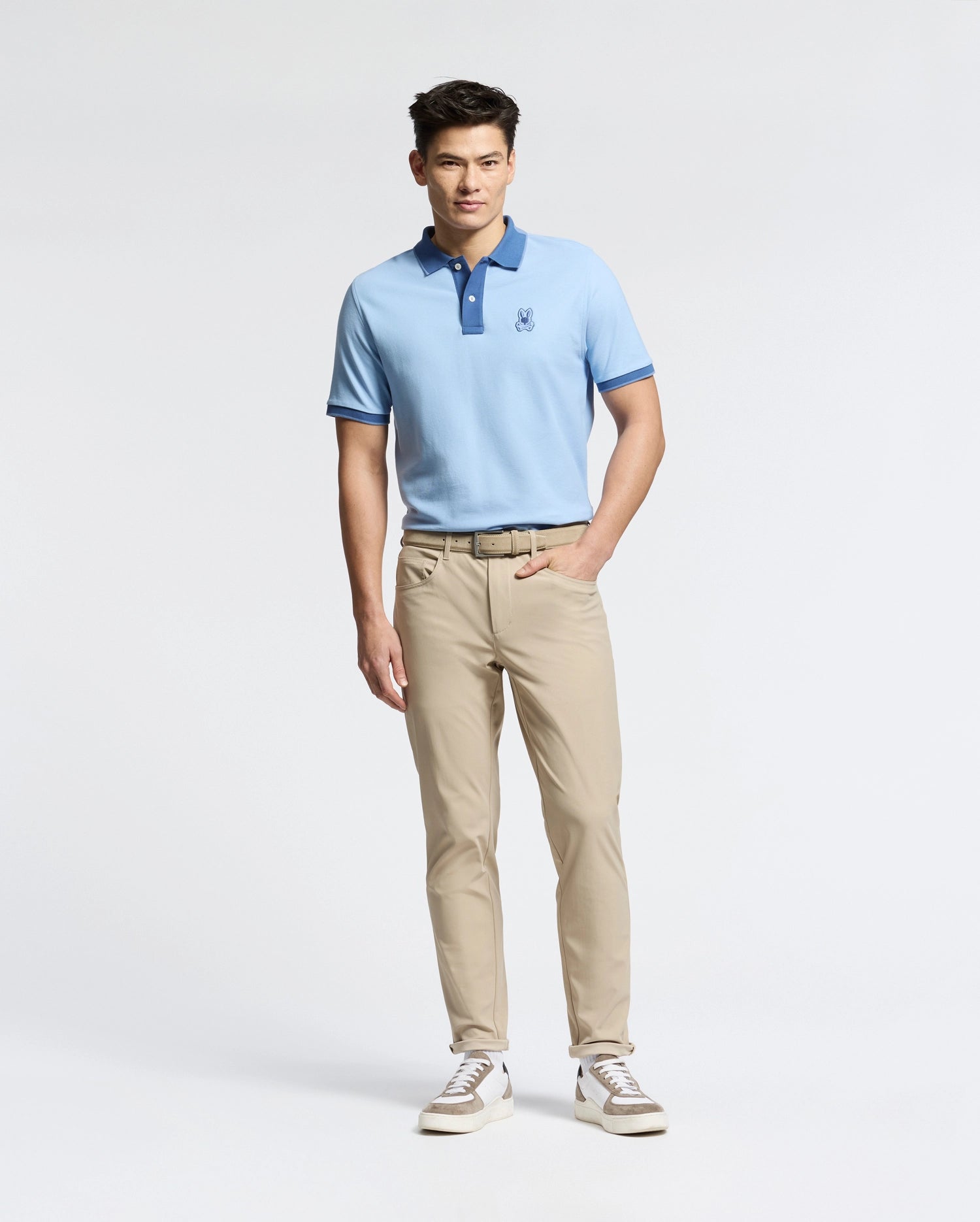 A man with short, dark hair stands against a plain background wearing the Psycho Bunny MENS NEW CASTLE PIQUE POLO - B6K487F200 in slim fit light blue with a darker blue collar and embroidered bunny patch. He pairs it with beige pants and white sneakers, his left hand in his pocket highlighting the Pima cotton fabric's elegance.