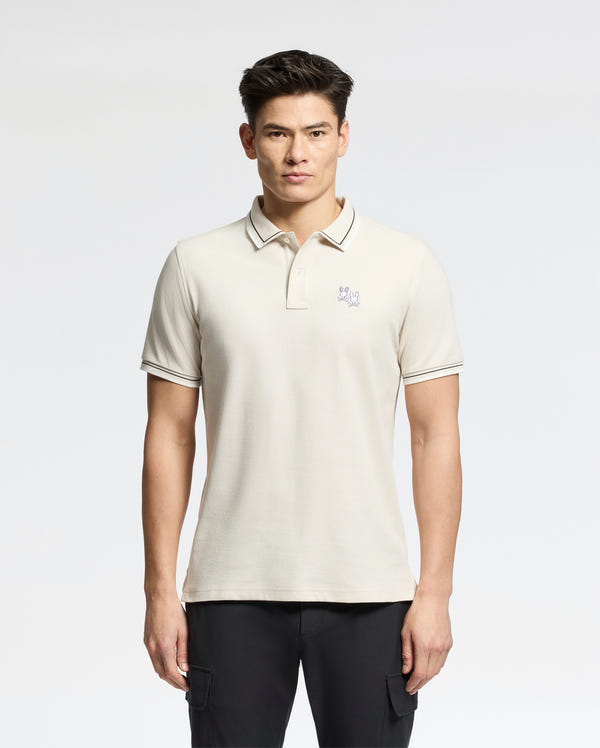 A person in a light-colored Psycho Bunny MENS ELLIOT PIQUE POLO - B6K505F200 made from soft Pima cotton with a small chest logo, paired with dark pants, stands against a white background.