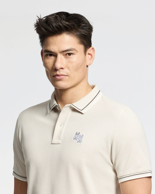 A person with short, dark hair is wearing a Psycho Bunny MENS ELLIOT PIQUE POLO. This Pima cotton shirt features a small bunny emblem on the chest, and they stand against a plain, light background with a neutral expression.