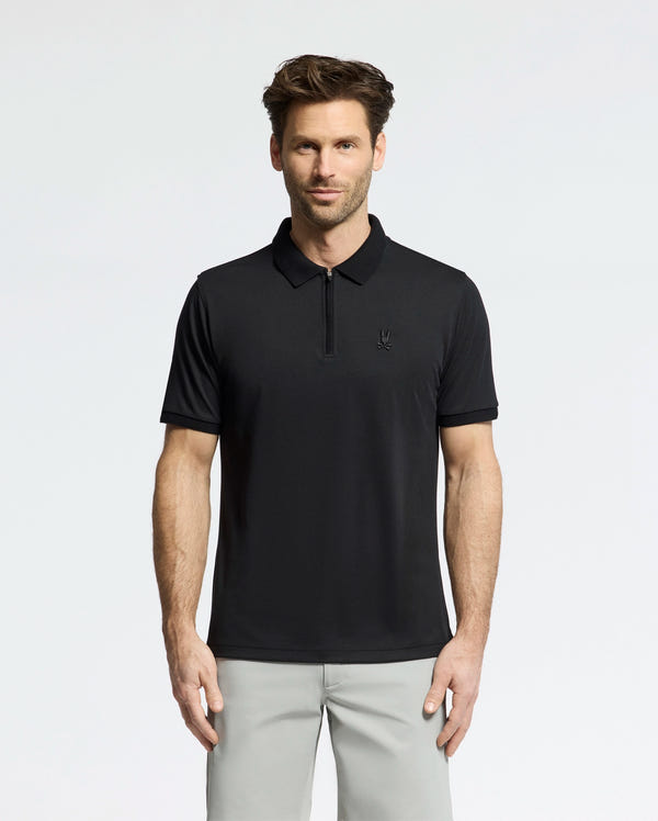 A man with short brown hair and a trimmed beard wears the Psycho Bunny MENS TREVOR PERFORMANCE PIQUE ZIP UP POLO (B6K557F200) in black and light gray pants, standing against a white background facing forward with a neutral expression.