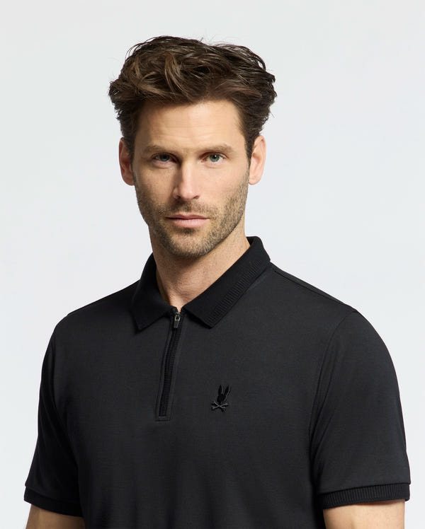 A man with short brown hair and light stubble wears the Psycho Bunny MENS TREVOR PERFORMANCE PIQUE ZIP UP POLO (B6K557F200), a slim fit black shirt with a small embroidered Bunny logo, looking directly at the camera against a plain light background.
