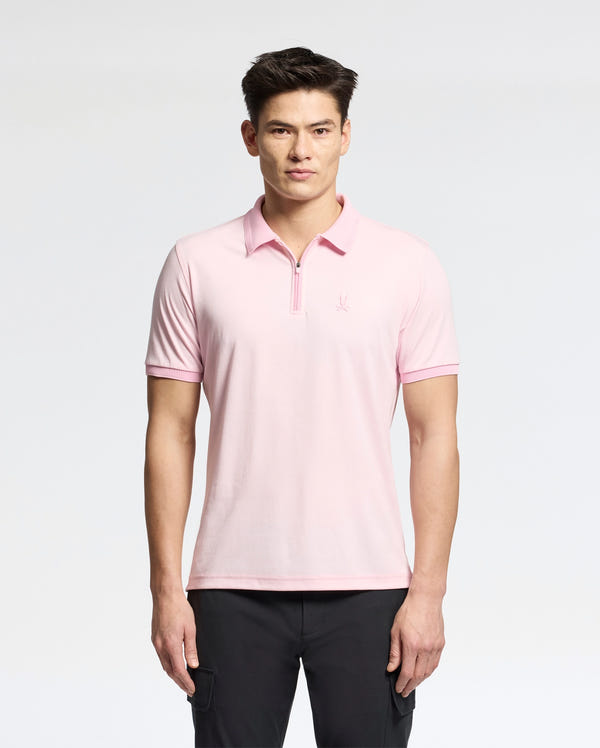 A man stands facing forward, wearing a light pink Psycho Bunny MENS TREVOR PERFORMANCE PIQUE ZIP UP POLO (B6K557F200) and black pants against a plain white background.