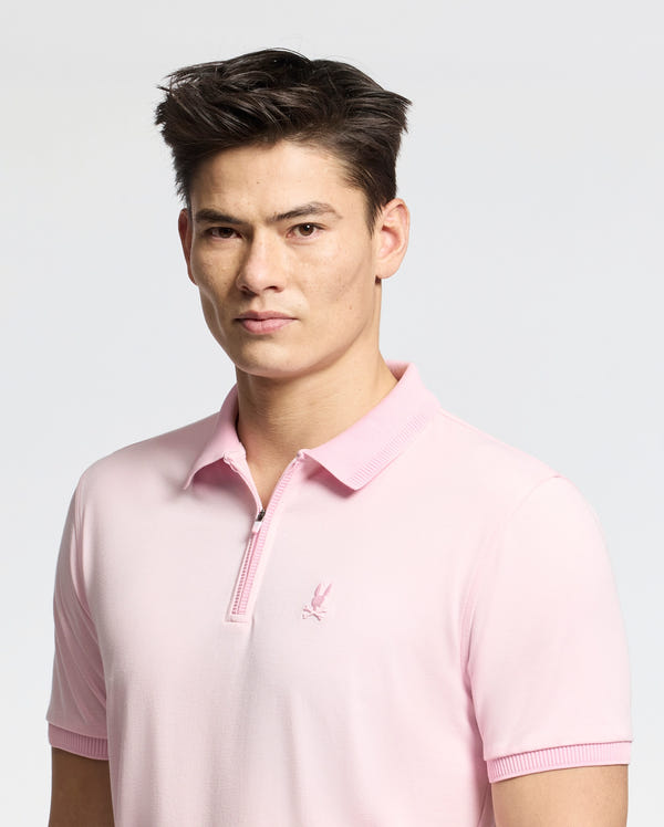 A young man with short dark hair wears a slim fit light pink Psycho Bunny MENS TREVOR PERFORMANCE PIQUE ZIP UP POLO (B6K557F200) featuring soft Pima cotton and a small embroidered logo, standing against a plain light background.