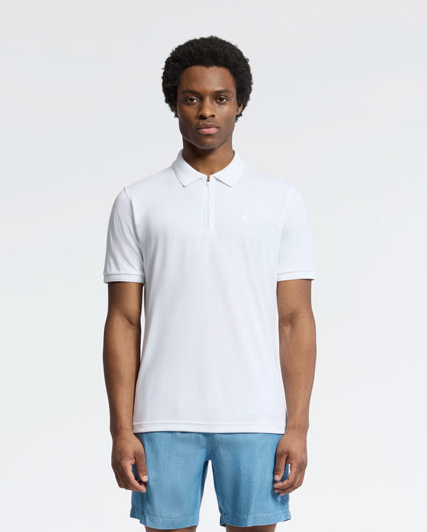 A man faces forward in a Psycho Bunny MENS TREVOR PERFORMANCE PIQUE ZIP UP POLO (B6K557F200), a slim fit white short-sleeve top, paired with light blue shorts, set against a plain white background.