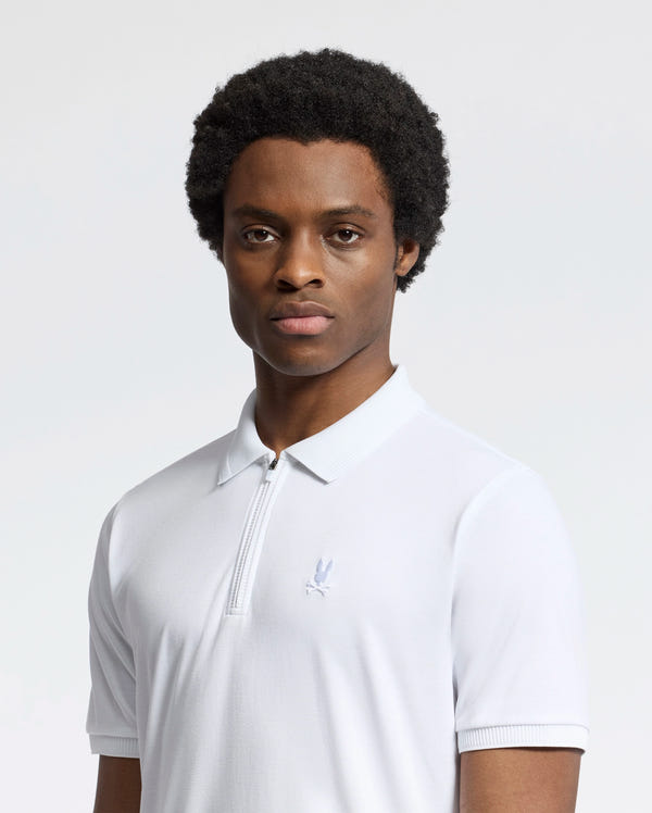 A man with short curly hair wears the Psycho Bunny MENS TREVOR PERFORMANCE PIQUE ZIP UP POLO (B6K557F200) in white, featuring a slim fit and chest emblem. He stands against a plain, light background, looking directly at the camera with a neutral expression.
