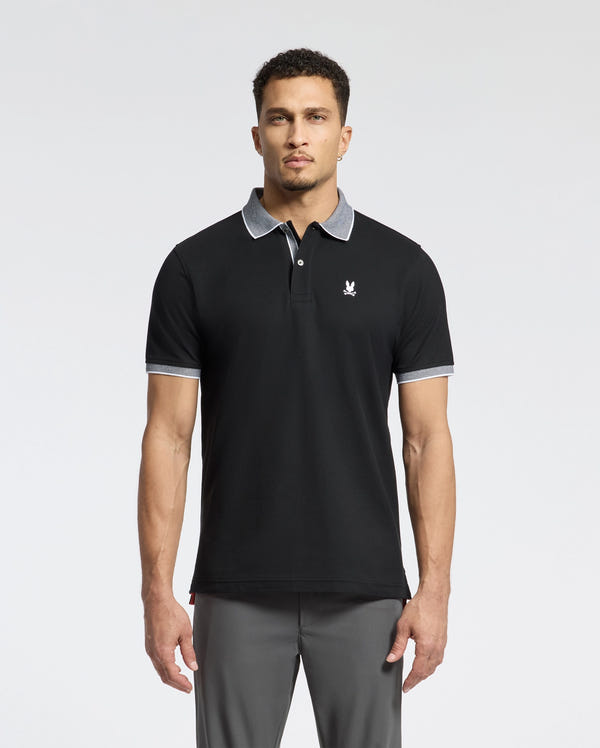 A man wears a Psycho Bunny MENS JONAS STRIPE COLLAR PIQUE POLO - B6K583F200. It's black, made from luxurious Pima Cotton with a gray collar and sleeve trim, featuring a small white emblem on the chest, showcasing its premium Peruvian quality against a plain white backdrop.