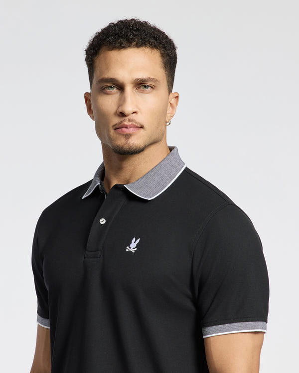 A man wears the Psycho Bunny MENS JONAS STRIPE COLLAR PIQUE POLO - B6K583F200 in black, featuring a gray collar and a small chest logo, made from luxurious Peruvian Pima Cotton, standing against a simple light background.