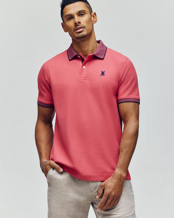 A man stands against a neutral background wearing beige pants and a Psycho Bunny MENS JONAS MICRO STRIPE COLLAR PIQUE POLO (B6K583F200) in coral pink, with one hand in his pocket.