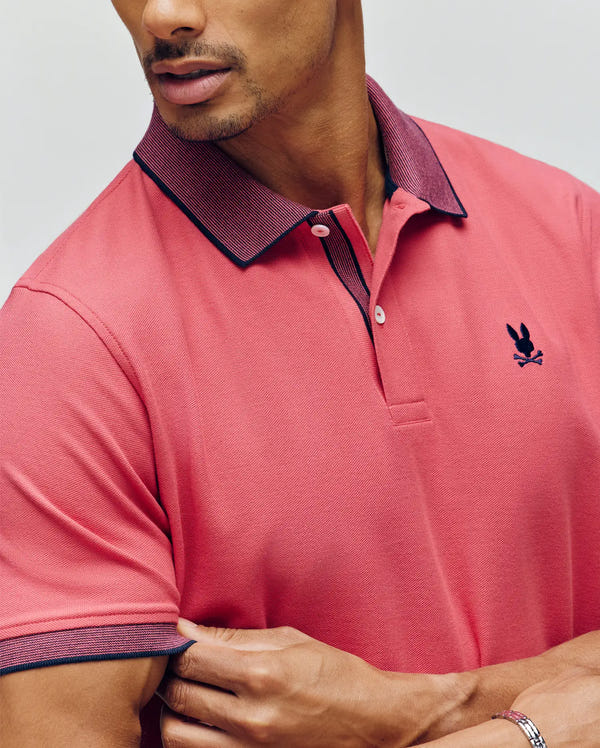 A man wearing the Psycho Bunny MENS JONAS MICRO STRIPE COLLAR PIQUE POLO (B6K583F200) with a pink body, dark collar and cuffs, embroidered Bunny logo on the chest, and micro-stripe details, crosses his arms against a light background.