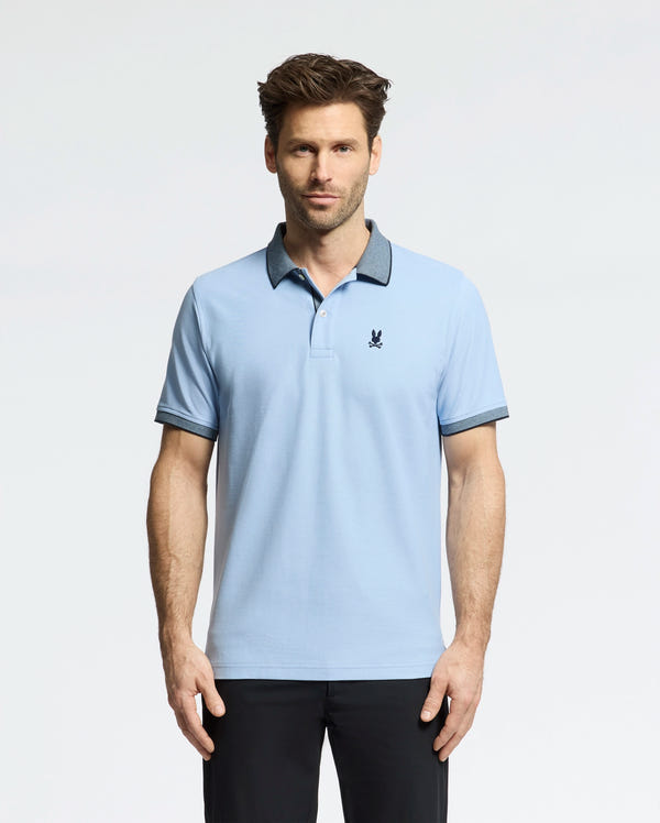 A man wears a light blue Psycho Bunny Men's Jonas Micro Stripe Collar Pique Polo, with a small dark emblem on the chest and micro-stripes, against a white backdrop. The shirt's dark collar and cuffs match his sleek black pants.