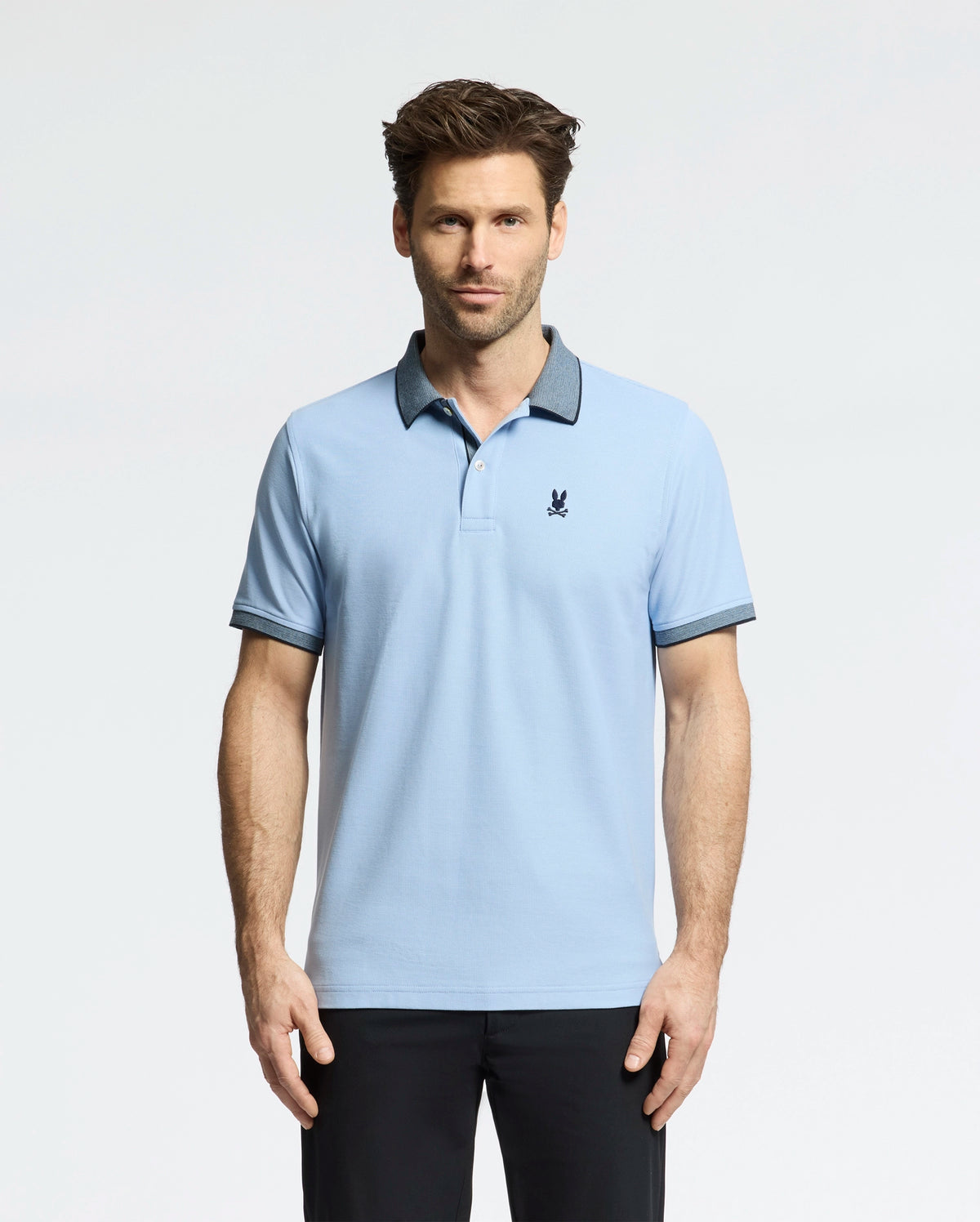 A man wears a light blue Psycho Bunny Men's Jonas Micro Stripe Collar Pique Polo, with a small dark emblem on the chest and micro-stripes, against a white backdrop. The shirt's dark collar and cuffs match his sleek black pants.