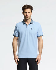 A man wears a light blue Psycho Bunny Men's Jonas Micro Stripe Collar Pique Polo, with a small dark emblem on the chest and micro-stripes, against a white backdrop. The shirt's dark collar and cuffs match his sleek black pants.