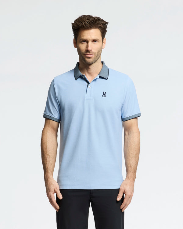 A man wears a light blue Psycho Bunny Men's Jonas Micro Stripe Collar Pique Polo, with a small dark emblem on the chest and micro-stripes, against a white backdrop. The shirt's dark collar and cuffs match his sleek black pants.