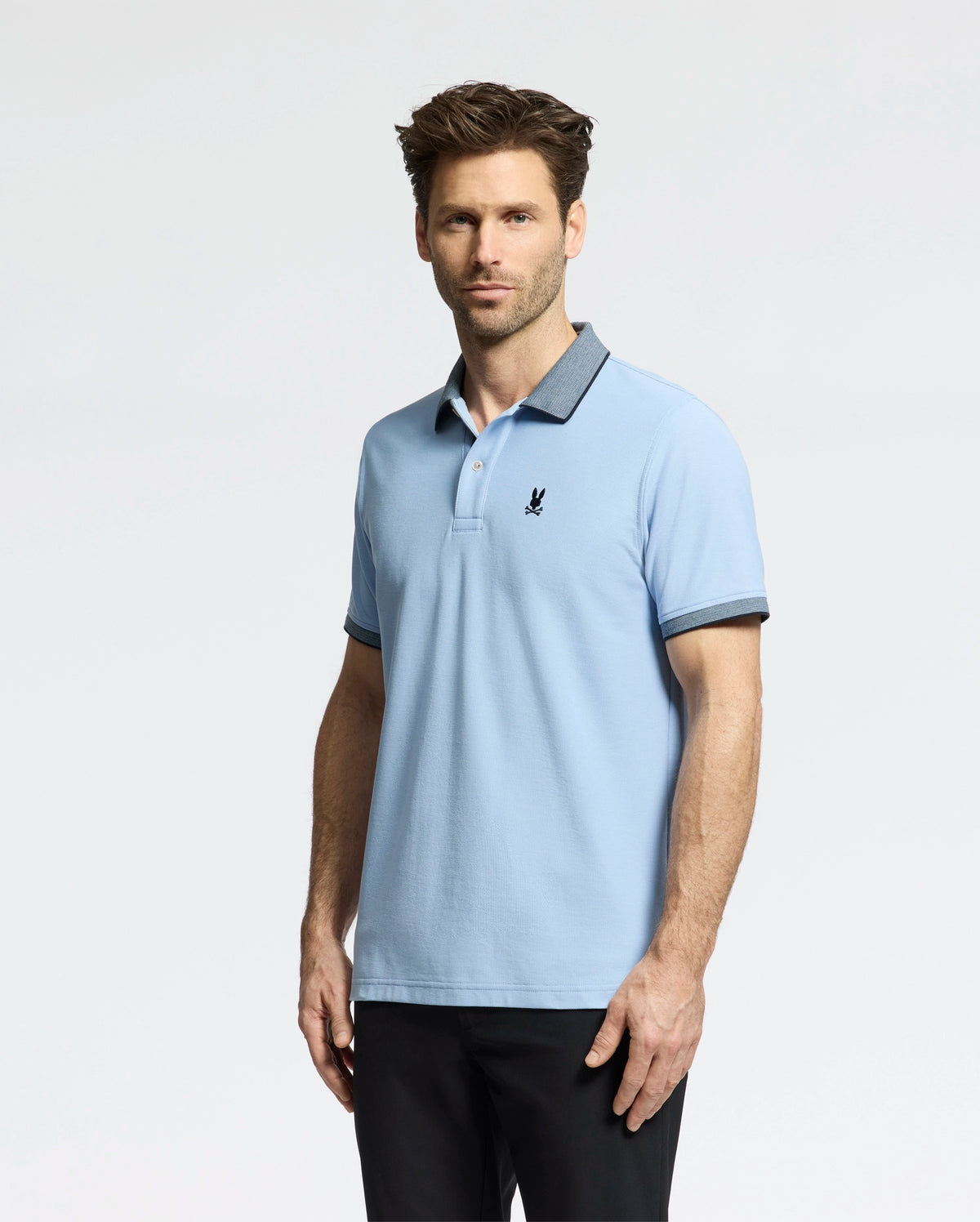 A man in a Psycho Bunny light blue MENS JONAS MICRO STRIPE COLLAR PIQUE POLO, marked with a black emblem on the left chest, and dark pants poses against a white background. The polo features micro-stripe detailing on its dark collar and sleeve trims. He has short brown hair and stubble.