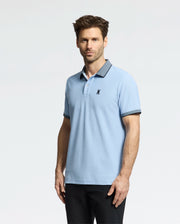 A man in a Psycho Bunny light blue MENS JONAS MICRO STRIPE COLLAR PIQUE POLO, marked with a black emblem on the left chest, and dark pants poses against a white background. The polo features micro-stripe detailing on its dark collar and sleeve trims. He has short brown hair and stubble.
