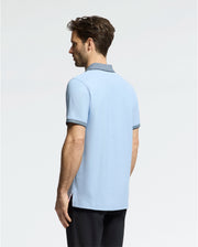 A man, with short brown hair, models a Psycho Bunny MENS JONAS MICRO STRIPE COLLAR PIQUE POLO in light blue with dark collar trim, made of premium Pima cotton. He pairs it with black pants against a plain, light-colored background.