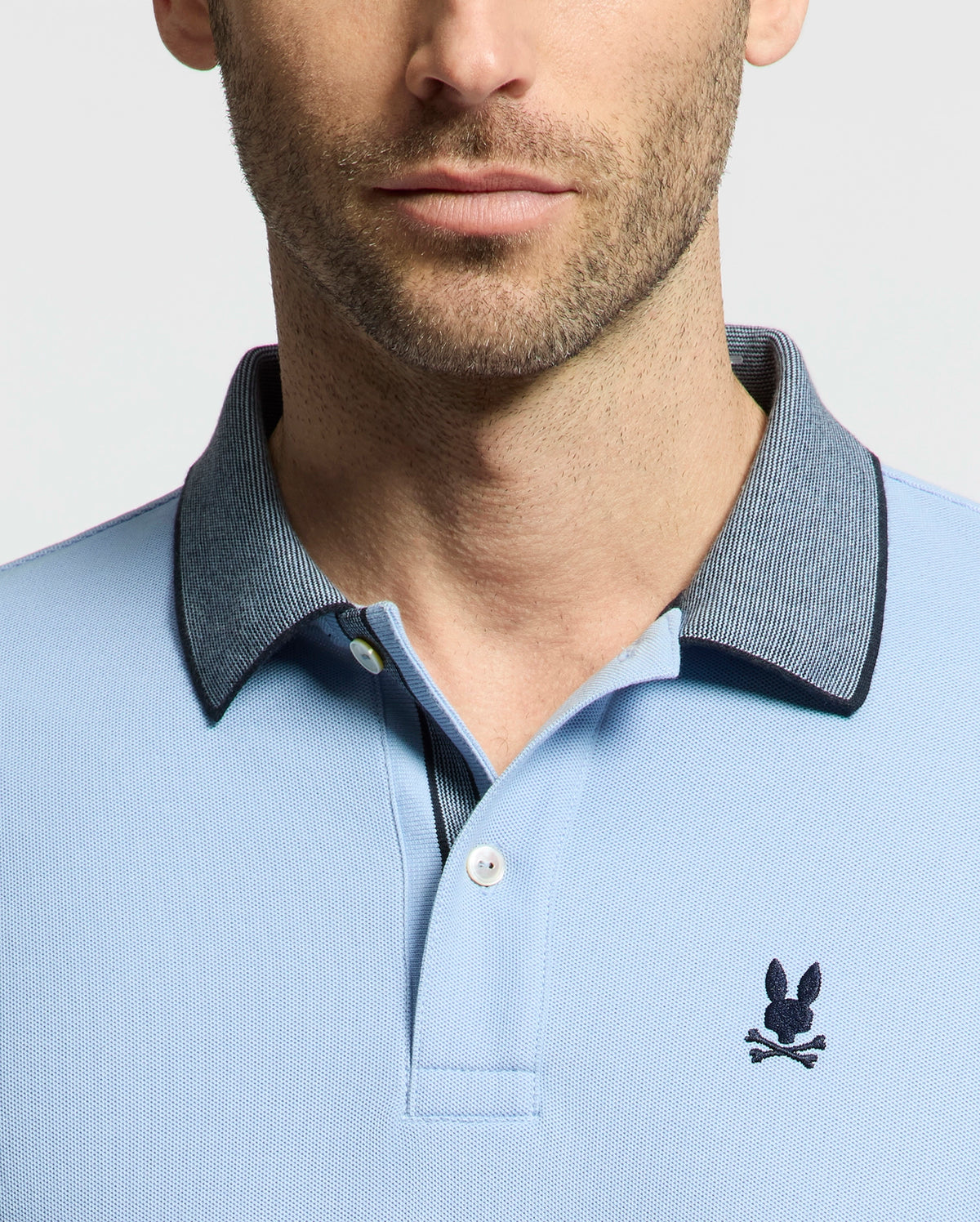 A man sports the Psycho Bunny Men's Jonas Micro Stripe Collar Pique Polo - B6K583F200 in light blue, made of soft Pima cotton with a gray collar. The polo showcases an embroidered rabbit head and crossed bones on the chest, accentuated by a plain light background that enhances its timeless micro-stripe pattern.