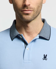 A man sports the Psycho Bunny Men's Jonas Micro Stripe Collar Pique Polo - B6K583F200 in light blue, made of soft Pima cotton with a gray collar. The polo showcases an embroidered rabbit head and crossed bones on the chest, accentuated by a plain light background that enhances its timeless micro-stripe pattern.