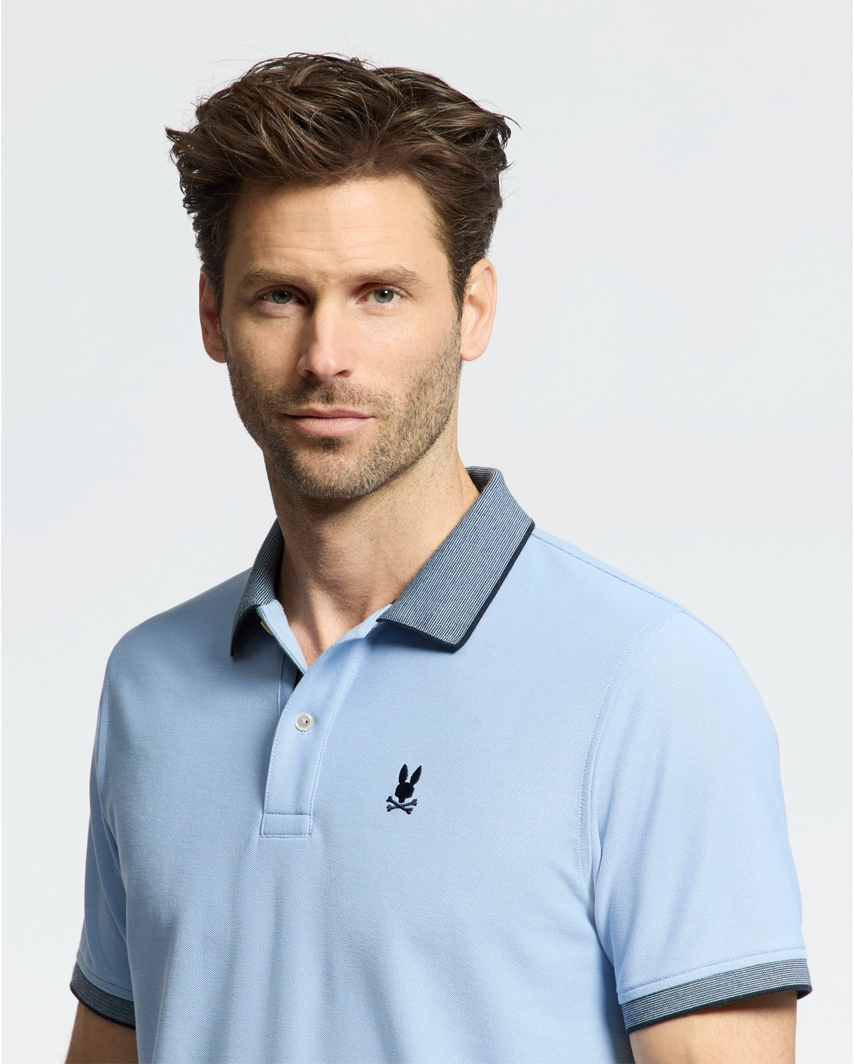 A man with brown hair and stubble wears a Psycho Bunny MENS JONAS MICRO STRIPE COLLAR PIQUE POLO (B6K583F200) in light blue, featuring a dark collar and a chest logo, against a plain white backdrop.
