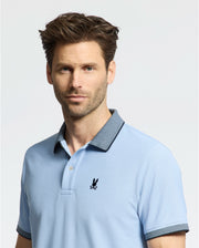 A man with brown hair and stubble wears a Psycho Bunny MENS JONAS MICRO STRIPE COLLAR PIQUE POLO (B6K583F200) in light blue, featuring a dark collar and a chest logo, against a plain white backdrop.