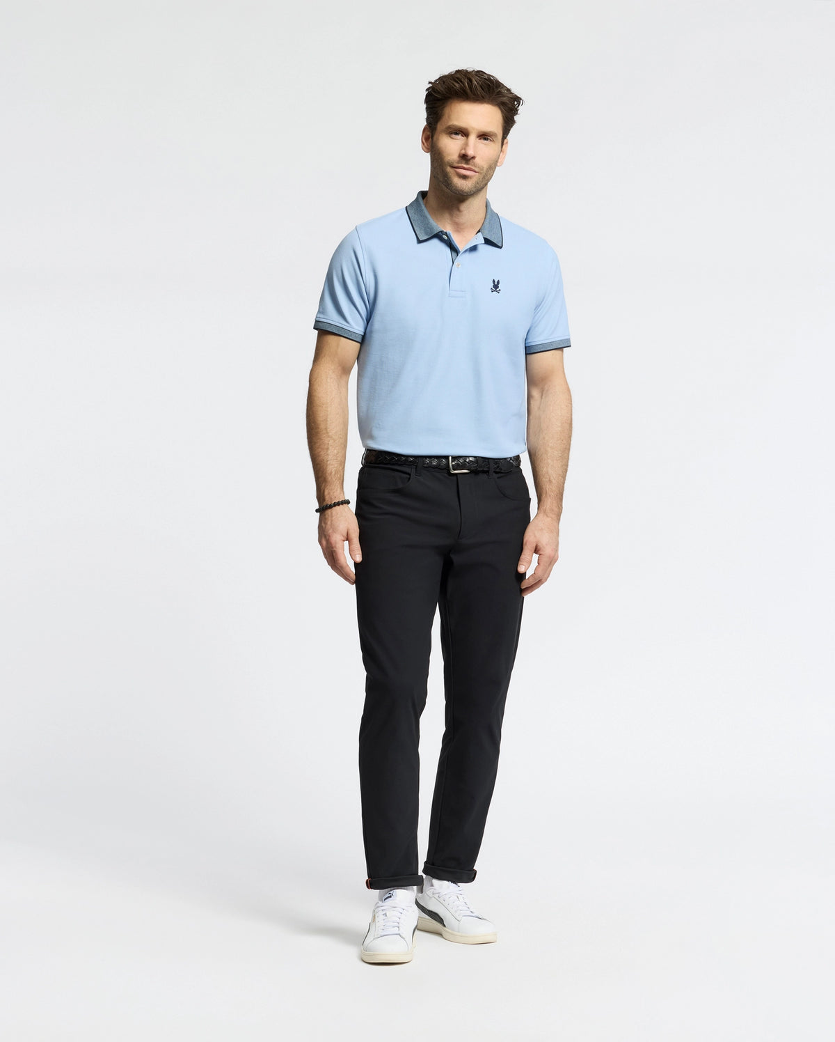 A man stands wearing a light blue Psycho Bunny MENS JONAS MICRO STRIPE COLLAR PIQUE POLO (B6K583F200) with black pants and white sneakers, featuring a micro-stripe pattern and chest logo. He has a slight beard, wears a bracelet on his left wrist, set against a plain white background.