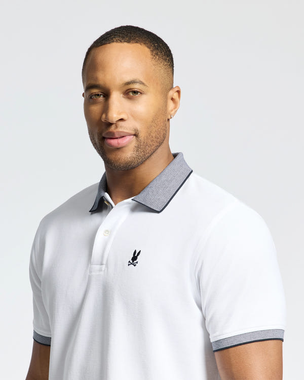 A person stands against a plain background wearing Psycho Bunny's MENS JONAS STRIPE COLLAR PIQUE POLO in white with a gray collar and an embroidered black logo on the chest. They have short hair and a subtly inviting smile.
