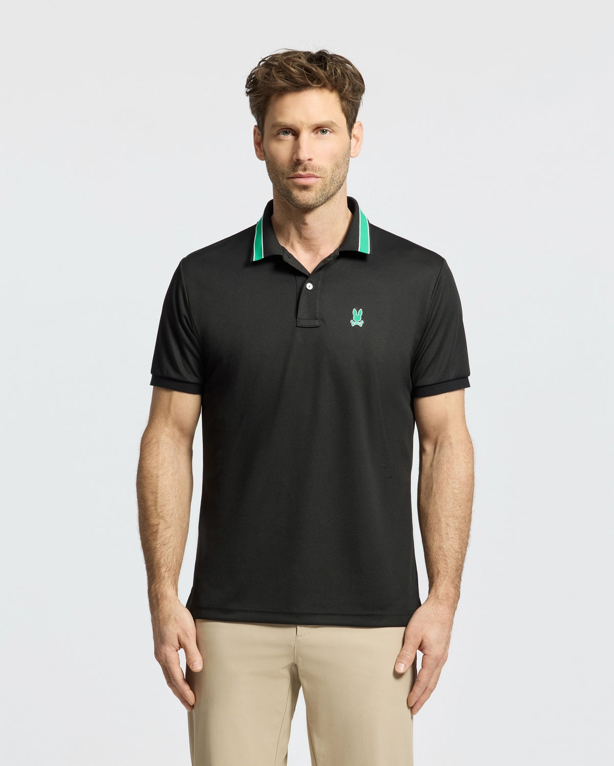 A man models the Psycho Bunny MENS FORREST SPORT PIQUE POLO (B6K660F200), featuring a slim-fit black design with green trim and chest logo, against a plain white backdrop. Paired with beige pants, he blends style and comfort seamlessly.