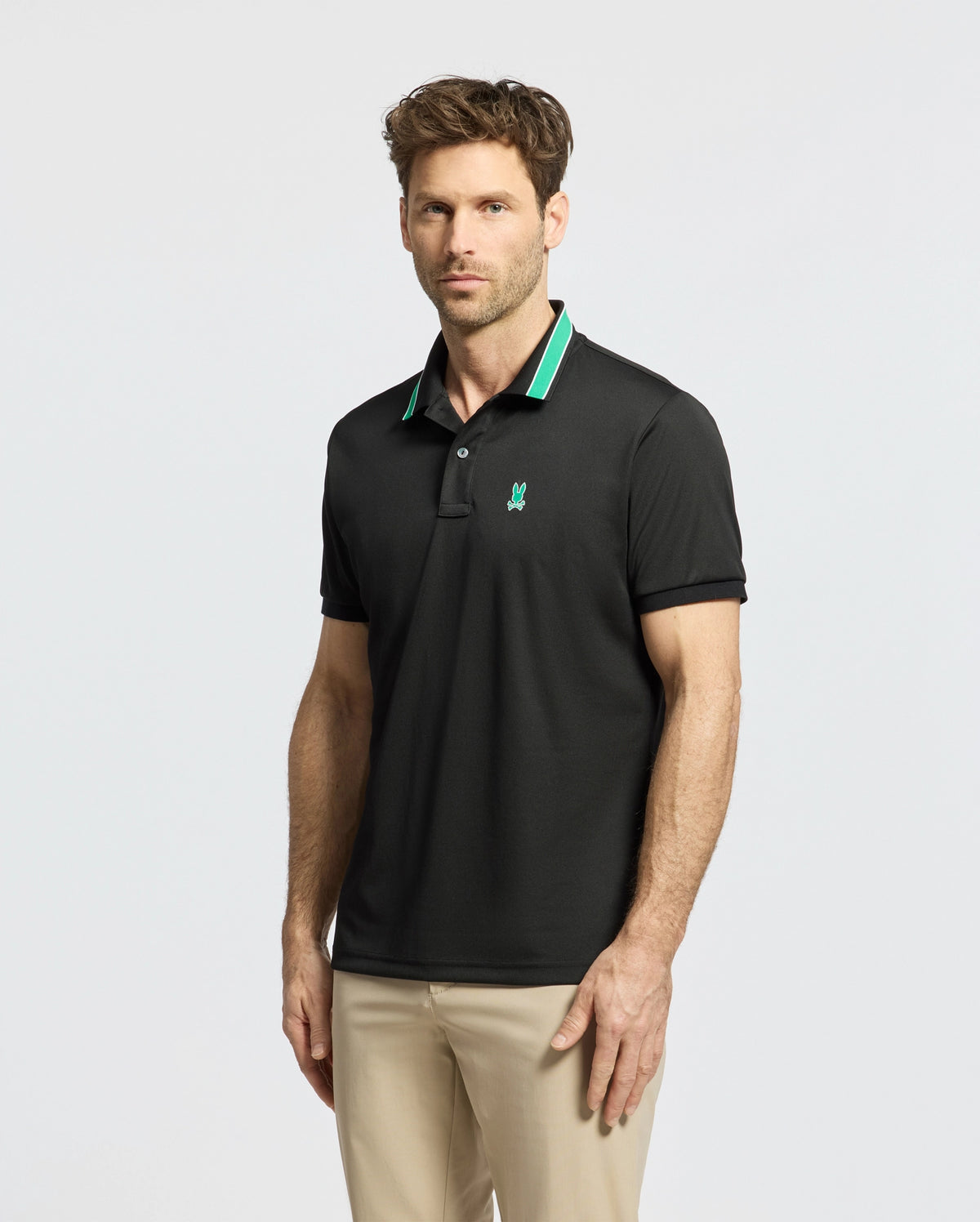 A man in a slim-fit Psycho Bunny MENS FORREST SPORT PIQUE POLO - B6K660F200, featuring black fabric with green accents, pairs it with beige pants. He stands against a plain white background, sporting short brown hair and a beard while looking directly at the camera.