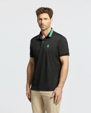 A man in a slim-fit Psycho Bunny MENS FORREST SPORT PIQUE POLO - B6K660F200, featuring black fabric with green accents, pairs it with beige pants. He stands against a plain white background, sporting short brown hair and a beard while looking directly at the camera.