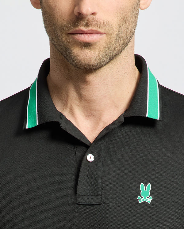 A man wears the MENS FORREST SPORT PIQUE POLO by Psycho Bunny, featuring a black body with green and white collar trim. This slim-fit shirt includes a small light blue rabbit and crossed bones logo on the left chest. Only his neck and lower face are visible.