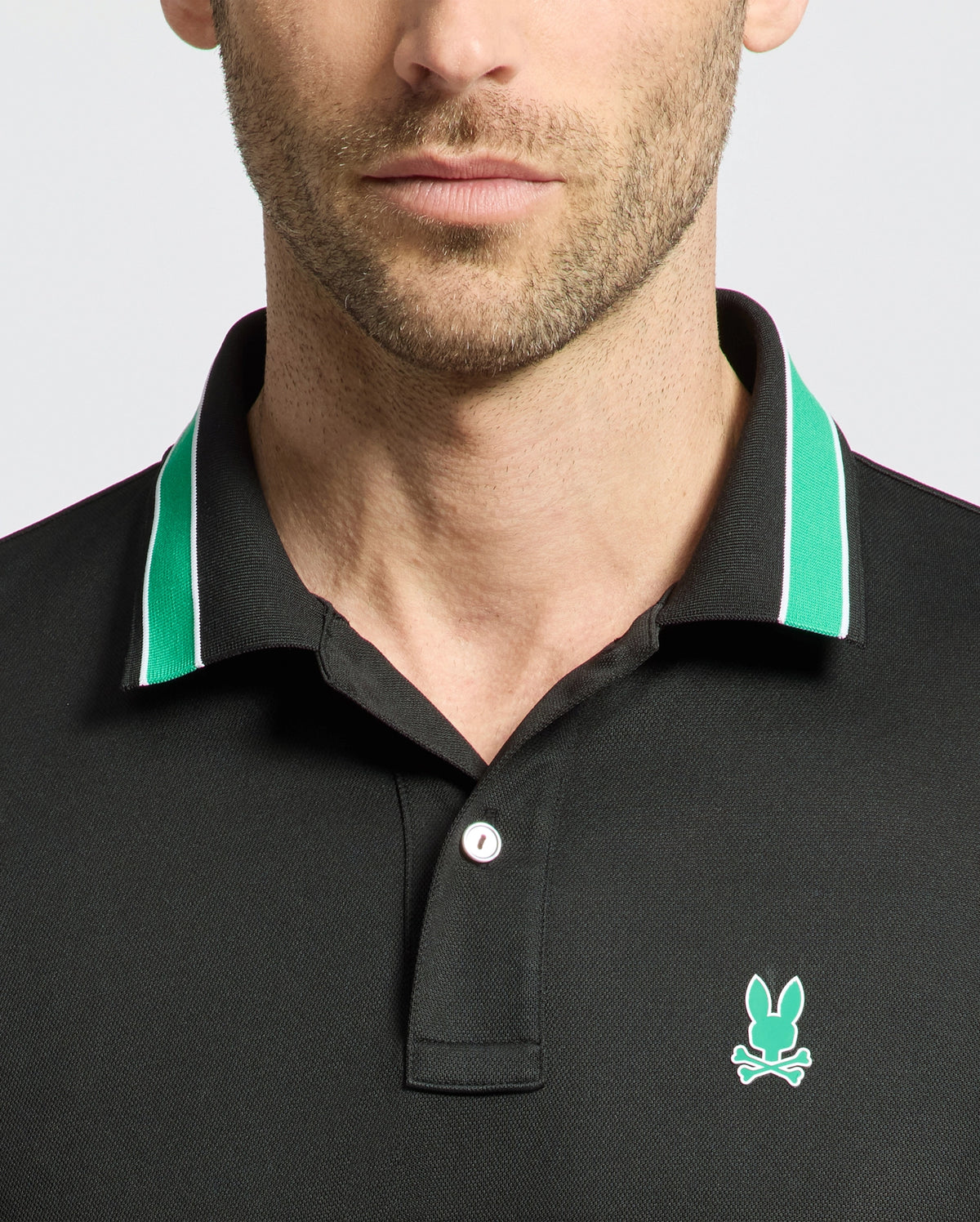 A man wears the MENS FORREST SPORT PIQUE POLO by Psycho Bunny, featuring a black body with green and white collar trim. This slim-fit shirt includes a small light blue rabbit and crossed bones logo on the left chest. Only his neck and lower face are visible.