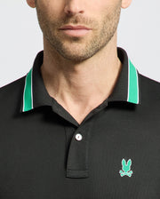 A man wears the MENS FORREST SPORT PIQUE POLO by Psycho Bunny, featuring a black body with green and white collar trim. This slim-fit shirt includes a small light blue rabbit and crossed bones logo on the left chest. Only his neck and lower face are visible.