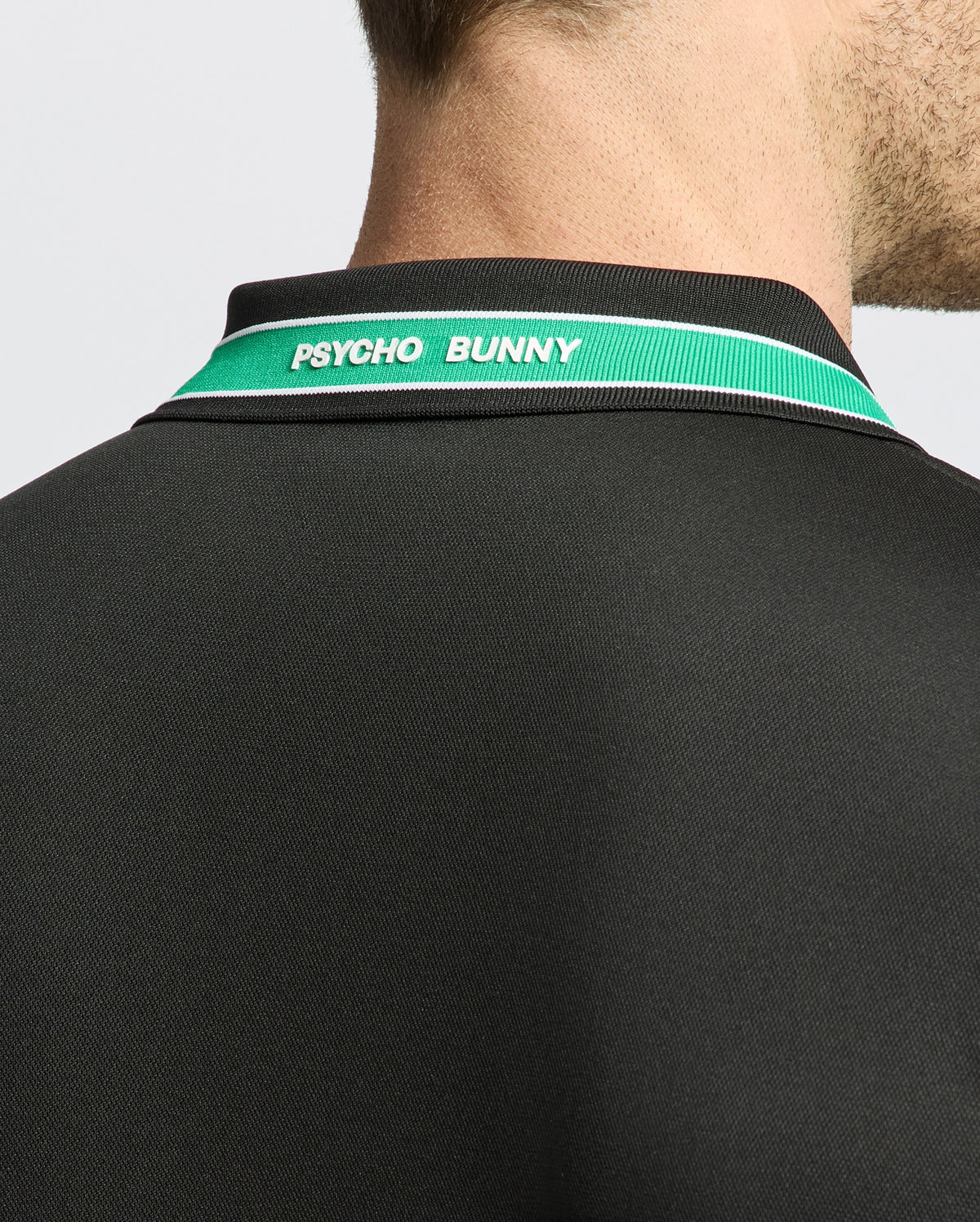Close-up of a person in the Psycho Bunny Forest Sport Piqué Polo, black with green and white collar accents. The back features "PSYCHO BUNNY" text. The shoulder, neck, and part of the head are visible against a light background, showcasing its moisture-wicking material.