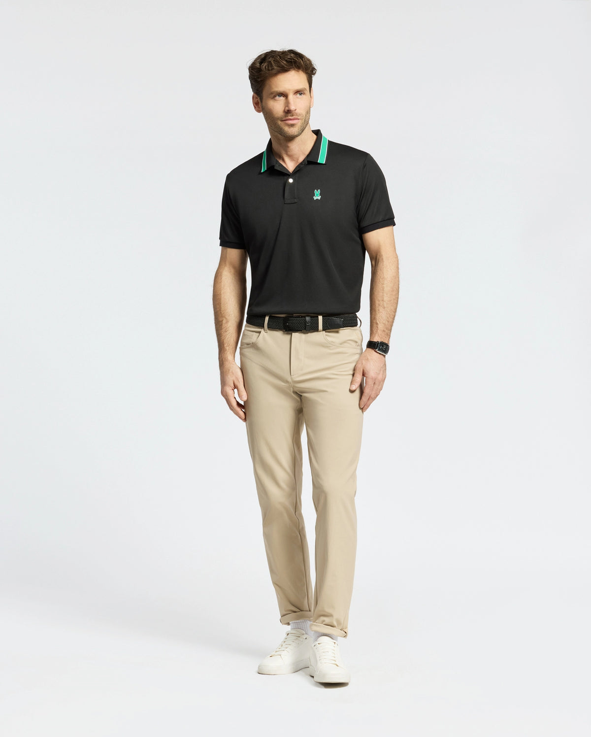 Wearing Psycho Bunny's MENS FORREST SPORT PIQUE POLO in slim-fit black with green accents, a man pairs it with beige pants and white sneakers. The moisture-wicking fabric ensures comfort against the plain white backdrop, and he completes his look with a black watch.