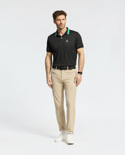 Wearing Psycho Bunny's MENS FORREST SPORT PIQUE POLO in slim-fit black with green accents, a man pairs it with beige pants and white sneakers. The moisture-wicking fabric ensures comfort against the plain white backdrop, and he completes his look with a black watch.