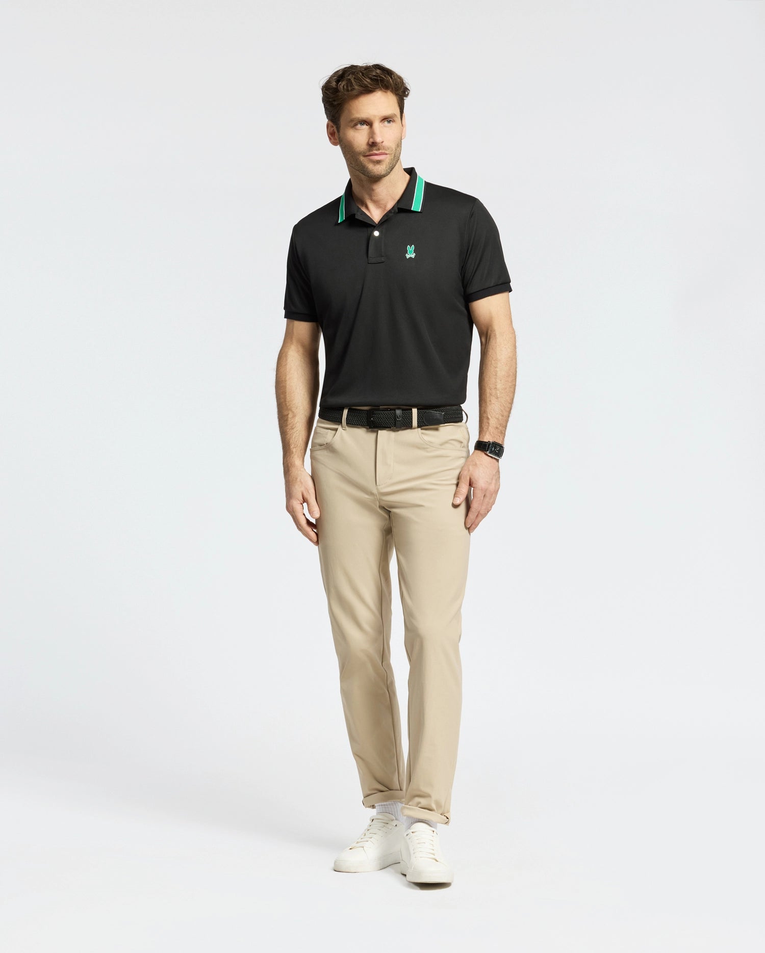Wearing Psycho Bunny's MENS FORREST SPORT PIQUE POLO in slim-fit black with green accents, a man pairs it with beige pants and white sneakers. The moisture-wicking fabric ensures comfort against the plain white backdrop, and he completes his look with a black watch.