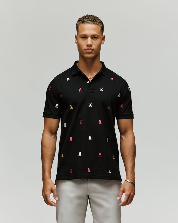 A man faces forward, wearing the Psycho Bunny MENS HOLLAND ALL OVER BUNNY PIQUE POLO (B6K618F200) in black with colorful embroidered bunny logos, paired with light grey pants against a plain, light background.