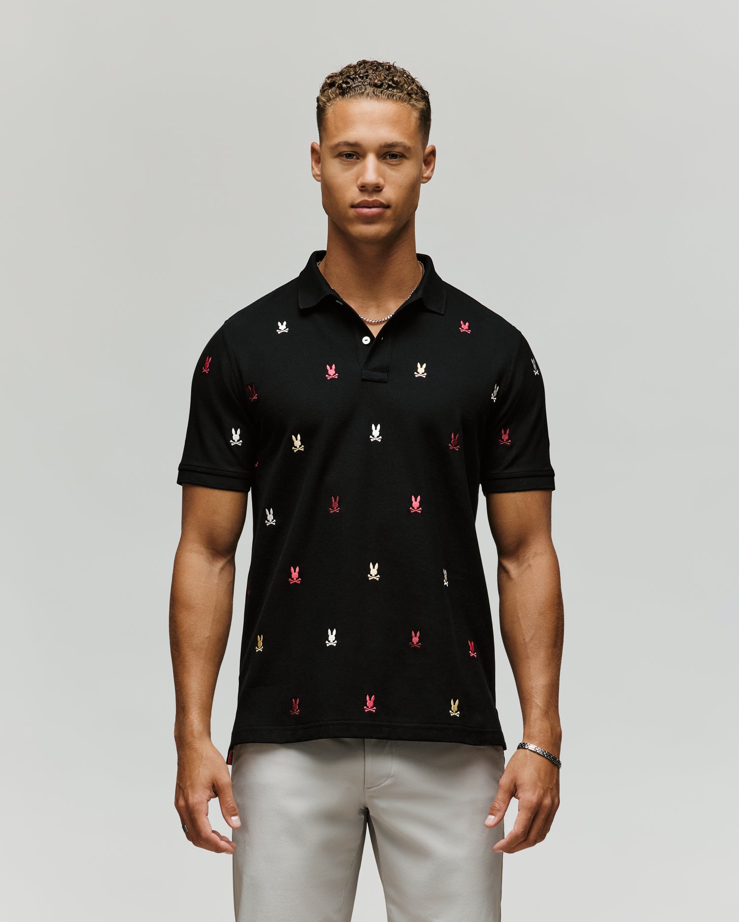 A man faces forward, wearing the Psycho Bunny MENS HOLLAND ALL OVER BUNNY PIQUE POLO (B6K618F200) in black with colorful embroidered bunny logos, paired with light grey pants against a plain, light background.