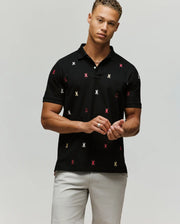 A man wears the Psycho Bunny MENS HOLLAND ALL OVER BUNNY PIQUE POLO (B6K618F200) in black, featuring colorful embroidered bunny logos, paired with light gray shorts, standing against a plain background looking at the camera.