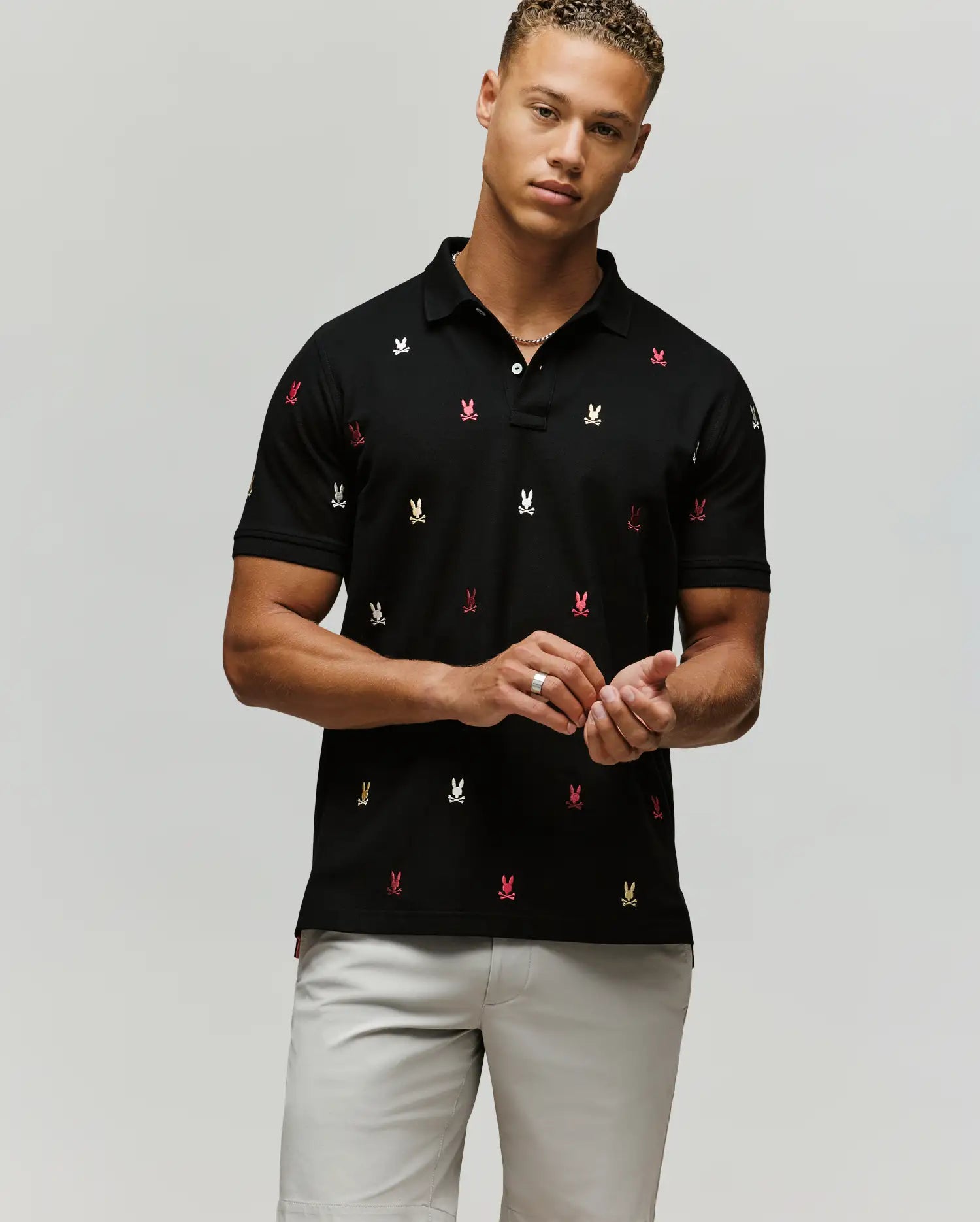 A man wears the Psycho Bunny MENS HOLLAND ALL OVER BUNNY PIQUE POLO (B6K618F200) in black, featuring colorful embroidered bunny logos, paired with light gray shorts, standing against a plain background looking at the camera.