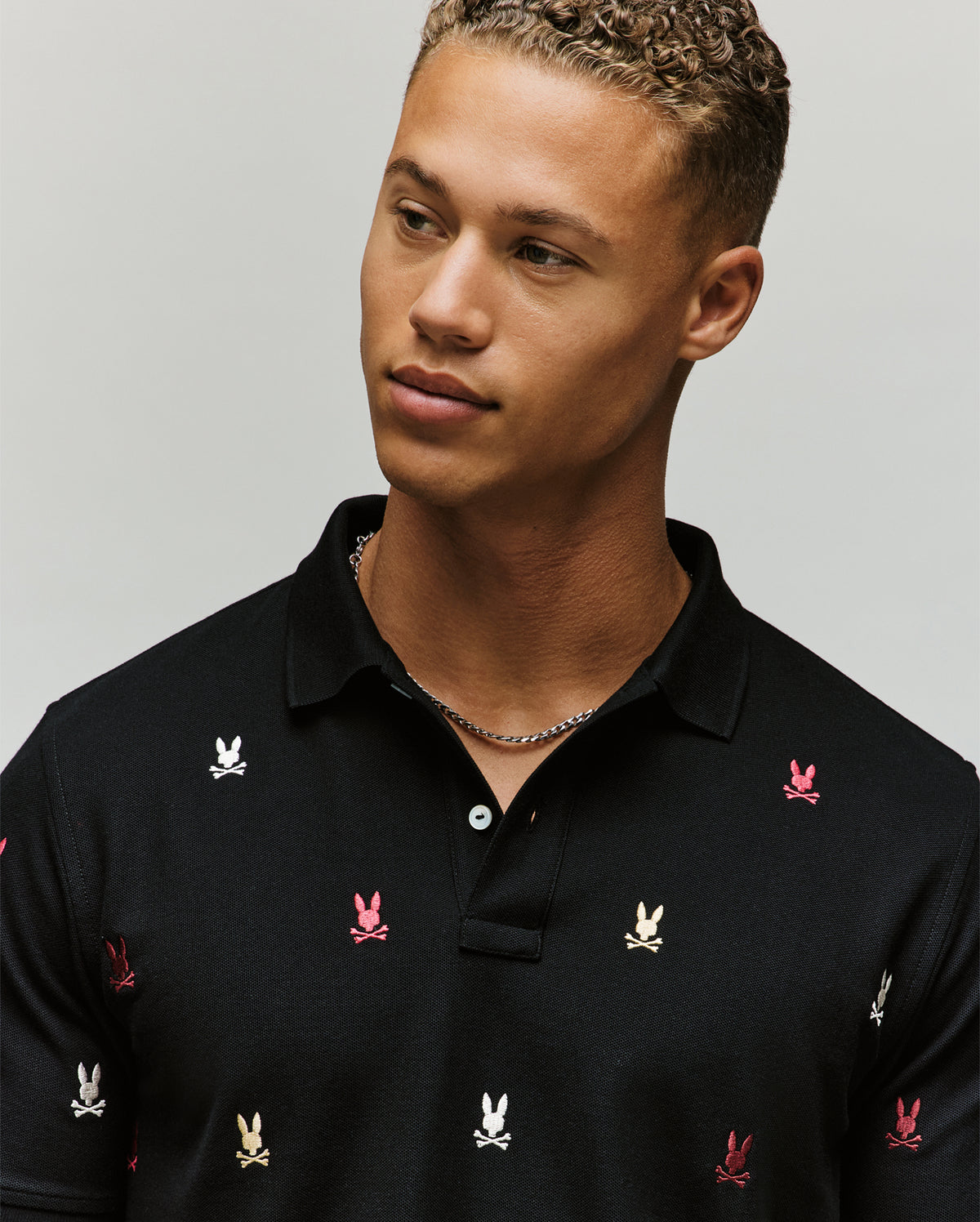 A young man with short curly hair wears a Psycho Bunny MENS HOLLAND ALL OVER BUNNY PIQUE POLO (B6K618F200) in black, featuring small white, red, and tan bunny and crossbones patterns. He looks to the side with a neutral expression and a silver chain necklace.