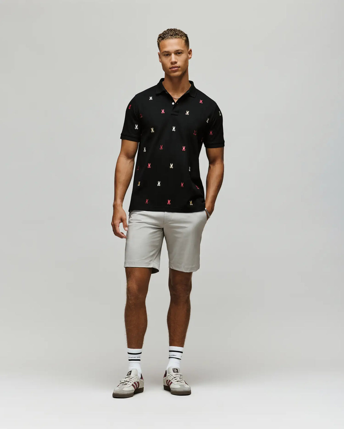 A man stands against a light gray background wearing the Psycho Bunny MENS HOLLAND ALL OVER BUNNY PIQUE POLO (B6K618F200), paired with light gray shorts, striped socks, and white sneakers. His hands rest relaxed at his sides.