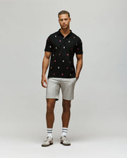 A man stands against a light gray background wearing the Psycho Bunny MENS HOLLAND ALL OVER BUNNY PIQUE POLO (B6K618F200), paired with light gray shorts, striped socks, and white sneakers. His hands rest relaxed at his sides.